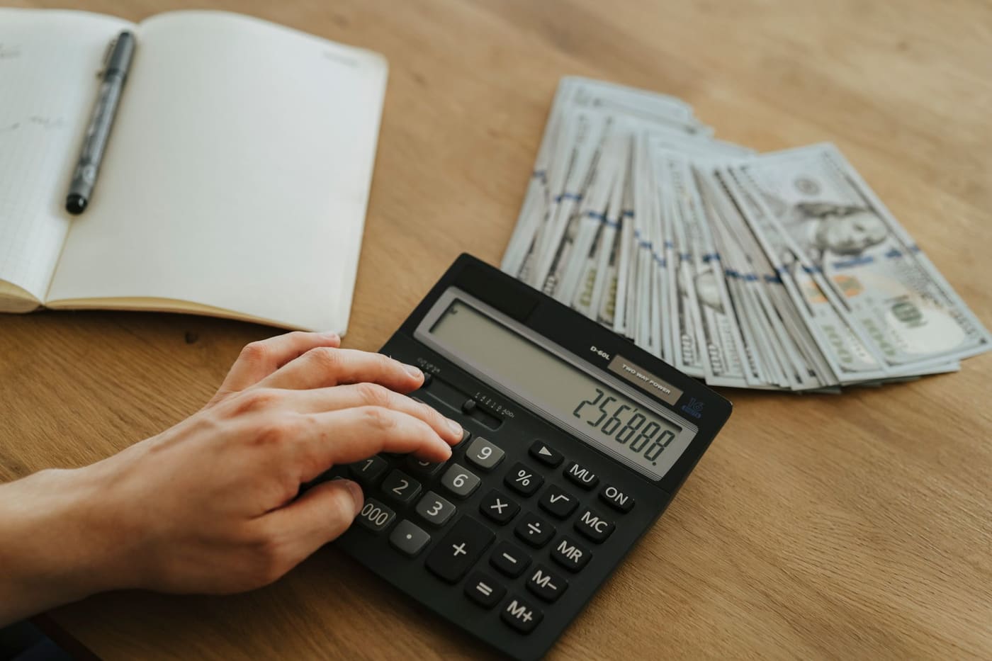 A hand using a calculator beside an open notebook on a wooden desk