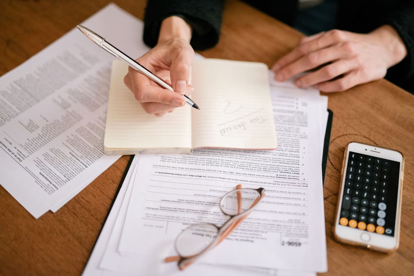 A hand writing in a notebook beside printed documents, glasses and a phone showing a calculator